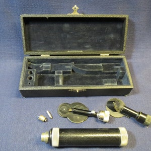 May include: A vintage black ophthalmoscope with a metal case. The ophthalmoscope has a black handle and a silver lens. The case is made of black leather and has a brass latch.