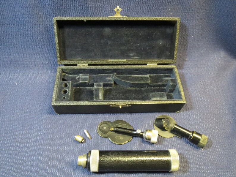 May include: A vintage black ophthalmoscope with a metal case. The ophthalmoscope has a black handle and a silver lens. The case is made of black leather and has a brass latch.