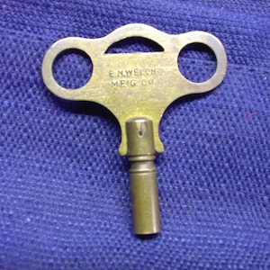 May include: A brass-colored metal clock winding key featuring a butterfly-shaped handle with two circular cutouts. The text E.N. WELCH MFG CO is embossed on the center of the handle. The key has a cylindrical shaft with a square-shaped opening at the base.