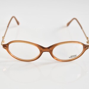 May include: Brown oval-shaped eyeglasses with gold accents and the text "Gianni Versace" on the right lens.