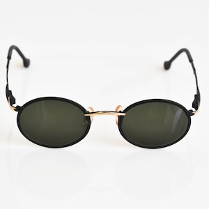 Puede incluir: Gafas de sol ovaladas negras con montura de metal dorado y lentes verde oscuro.