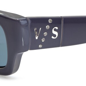 May include: Close-up of a pair of dark gray sunglasses with blue lenses. The sunglasses have a silver V:S logo with rhinestones on the side.