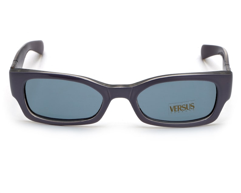 May include: Black rectangular sunglasses with dark blue lenses. The sunglasses have the word "VERSUS" printed on the right lens.