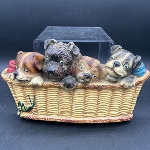 May include: A ceramic figurine of four dogs peeking out of a wicker basket. The dogs are brown, black, and white with painted details. The basket is brown and has a green grasshopper on the side.