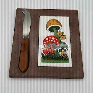 May include: A brown wooden cheese board with a decorative ceramic tile featuring colourful mushrooms and flowers. A cheese knife with a wooden handle and a curved blade is included. The board is a square shape.