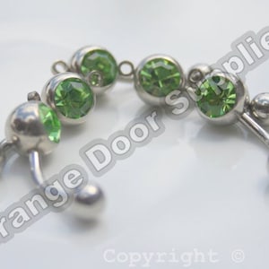 Op de afbeelding: Twee zilveren lichaamsieraden met groene edelstenen. Elk stuk heeft een gebogen staaf met twee ballen aan elk uiteinde.