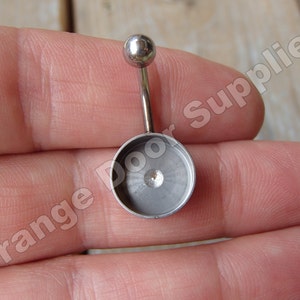 Puede incluir: Un piercing para el ombligo de color plateado con una pequeña bola en la parte superior y una base circular. La base tiene un pequeño perno centrado. Las palabras "Orange Door Supplies" son visibles en el fondo.