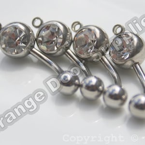 Puede incluir: Cuatro anillos de ombligo plateados con piedras de cristal transparentes. Cada anillo tiene una bola pequeña en la parte inferior y una bola más grande en la parte superior con una piedra de cristal.