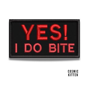 Puede incluir: Parche rectangular negro con las palabras "YES! I DO BITE" bordadas en hilo rojo. El parche tiene un borde negro y está sobre un fondo blanco. Las palabras están en una fuente en negrita sin serifa.
