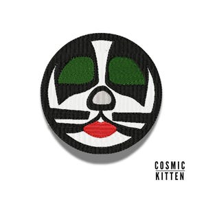 Pode incluir: Um remendo redondo bordado em preto e branco com um rosto estilizado. O rosto tem marcas pretas e brancas, olhos verdes e uma boca vermelha.