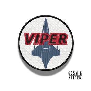 Puede incluir: Un parche circular blanco con borde negro. El parche presenta una nave espacial azul y gris con la palabra "VIPER" en letras rojas.