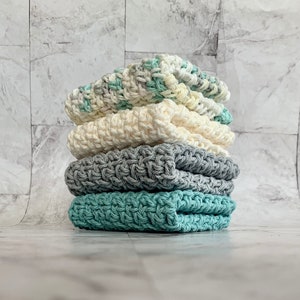 May include: A stack of four crocheted dishcloths in various colors, including green, gray, and white. The dishcloths are folded and stacked on top of each other.