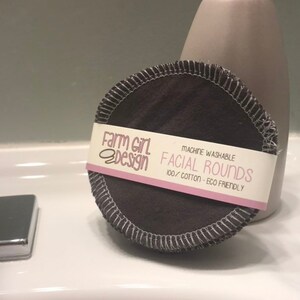 May include: A stack of dark gray reusable cotton facial rounds with a pink and white label that reads "Farm Girl Design" and "Machine Washable Facial Rounds 100% Cotton - Eco Friendly".