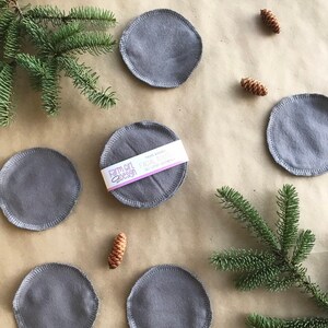May include: Six gray reusable makeup remover pads with a white label that reads "Farm Girl Design" and "Make-up Remover Pads". The pads are arranged in a circle on a brown paper background with pine branches and pine cones.