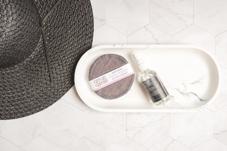 May include: A white oval tray with a gray reusable facial round and a clear glass bottle with a black label. The label reads "Farm Girl Design" and "Facial Rounds". The tray is on a white hexagonal tile surface.