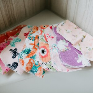 以下が含まれることがあります： フラミンゴ、蝶、ユニコーンなど、さまざまなパターンが施されたカラフルな布製パッドのセット。パッドは白いトリムで縫製されており、柔らかく吸収性のある質感です。