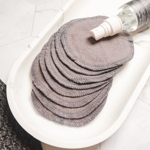 May include: A white ceramic tray with a stack of gray reusable makeup remover rounds and a clear spray bottle with a white cap.