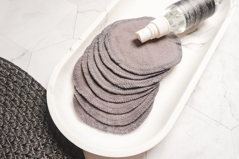 May include: A white ceramic tray with a stack of gray reusable makeup remover rounds and a clear spray bottle with a white cap.