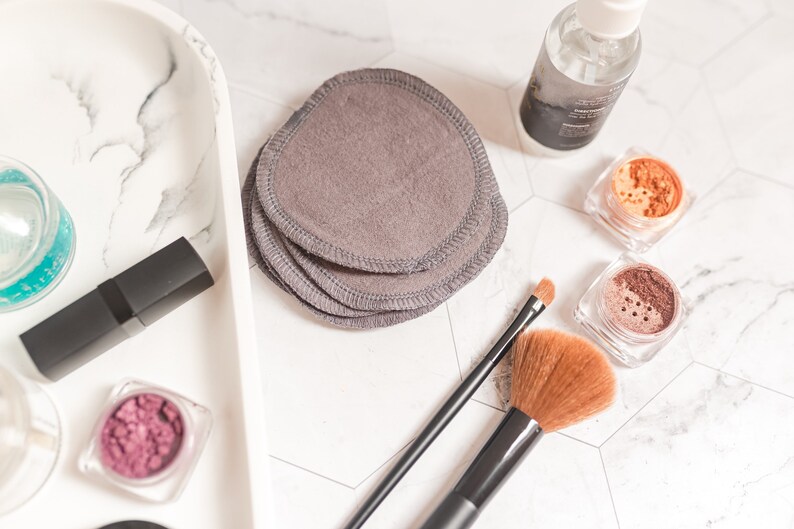May include: A white marble tray with a variety of makeup products, including a lipstick, eyeshadow, and a bottle of makeup remover. There are also a set of gray reusable makeup remover pads and two makeup brushes.