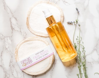 May include: A set of reusable, washable cotton facial rounds in a stack with a pink and white label that reads "Farm Girl Design, Made Washable, Facial Rounds, 100% Cotton, Eco Friendly". A clear glass bottle of facial oil with a gold pump top is next to the rounds on a white marble surface.