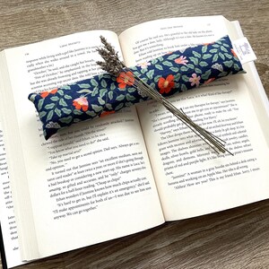 May include: An open book with a navy blue floral print lavender sachet and dried lavender sprigs. The sachet has a pattern of red and pink flowers. The book's pages are open, displaying text.