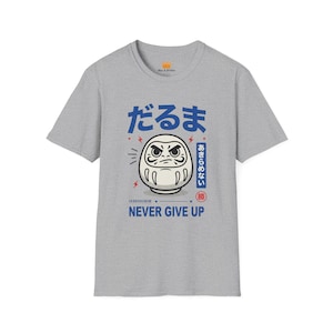 May include: Gray t-shirt featuring a Daruma doll illustration with Japanese text and the words "NEVER GIVE UP" in blue. The Daruma doll has a stern expression and is surrounded by lightning bolt graphics.