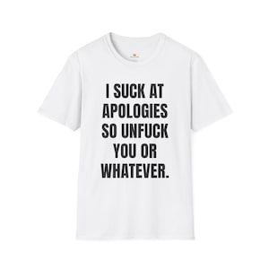 May include: White t-shirt with the text "I SUCK AT APOLOGIES SO UNFUCK YOU OR WHATEVER" in black, centered on the front. The shirt has a crew neck and short sleeves. A small gold crown is visible near the neckline.