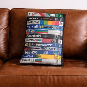 May include: A rectangular cushion displaying a collage of retro VHS tape labels, including brands such as Sony, Polaroid, and Maxell. The cushion is positioned on a brown leather sofa, creating a nostalgic feel.