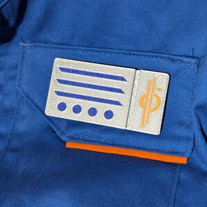 May include: A silver and blue patch with four white dots and a vertical orange stripe. The patch has a blue and white striped design and a vertical orange stripe with a stylized symbol.