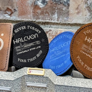 May include: Three leather coasters with the text "You Never Forget Halcyon Chandrila Star Line Your First Ship" in white lettering. The coasters are brown, black, and blue.
