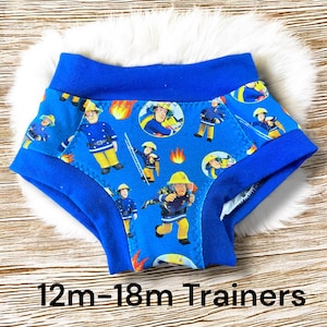 May include: Blue training pants with a Fireman Sam print. The pants have a blue elastic waistband and are labeled "12m-18m Trainers".