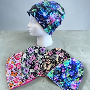 May include: A collection of five beanies featuring vibrant floral and butterfly patterns. One beanie is displayed on a white mannequin head, while four others are arranged in a row, showcasing designs with pink, purple, blue, and green colours.