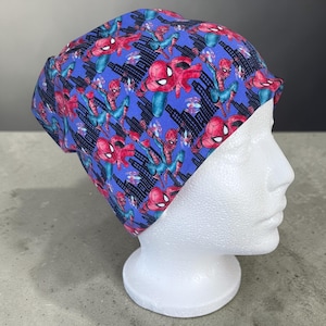 May include: A blue and red Spider-Man patterned beanie hat. The hat features a repeating design of the superhero in action poses, set against a cityscape backdrop. The hat is displayed on a white mannequin head.