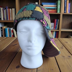 May include: A patchwork-patterned hat with a brim, displayed on a white mannequin head. The hat features teal, yellow, pink, and grey patches. The brim is grey, and the interior is light brown. The hat is a unique accessory.