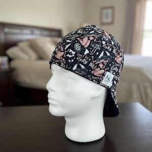 May include: Black welding cap with a repeating pattern of pin-up style illustrations in pink and white on a black background. The cap features the text "Don't be a Salty Bitch" and other phrases. A white label with the initials "LK" is visible.