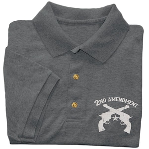 May include: A charcoal grey short-sleeve polo shirt featuring a white graphic print on the lower right side. The design displays two crossed revolvers with a small star in the center and the text 2ND AMENDMENT arched above the firearms.