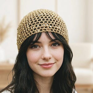 May include: A gold-colored, crocheted beanie hat with a net-like design and small sequins. The hat is worn by a person with dark hair and bangs. The background is a soft, neutral color.