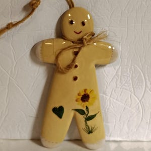 May include: A ceramic gingerbread man ornament with a yellow glaze. The ornament features a painted sunflower and a green heart. It has a jute string for hanging and is suitable for holiday decor.