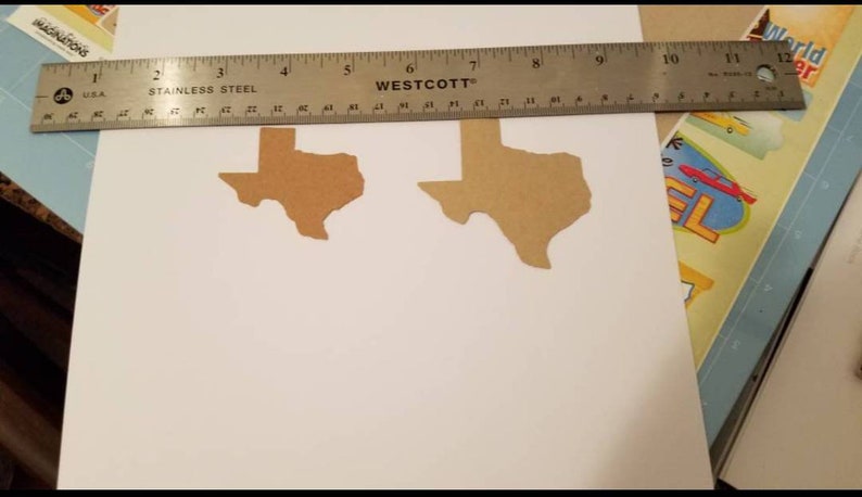 May include: Two brown paper cutouts of the state of Texas are on a white surface. A stainless steel ruler, marked in inches, is placed over the cutouts. The ruler is marked with the "Westcott" brand.