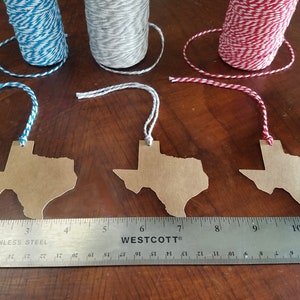 May include: Three brown paper Texas-shaped tags with twine hangers, next to spools of blue, gray, and red and white twine. A stainless steel ruler is placed below the tags, measuring in inches.