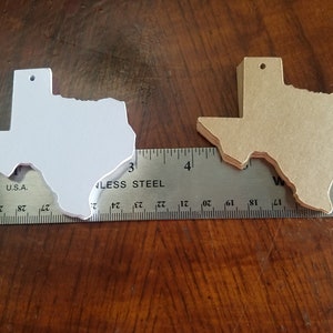May include: Two Texas-shaped cutouts, one white and one tan, are displayed next to a ruler. The white cutout is on the left, and the tan cutout is on the right. The ruler shows measurements in inches.