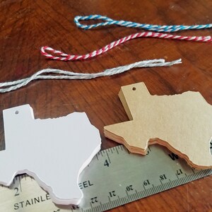 May include: Stacks of white and tan Texas-shaped tags with pre-drilled holes, alongside colorful twine and a stainless steel ruler. The tags are stacked on a wooden surface, with the ruler providing a size reference.