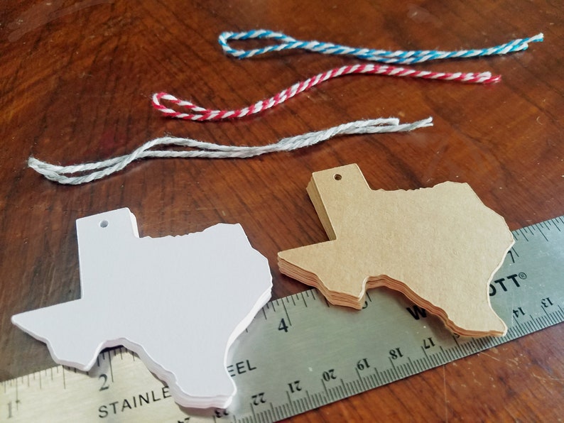 May include: Stacks of white and tan Texas-shaped tags with pre-drilled holes, alongside colorful twine and a stainless steel ruler. The tags are stacked on a wooden surface, with the ruler providing a size reference.