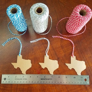 May include: Three spools of twine in blue and white, gray and white, and red and white, with matching Texas-shaped tags. A stainless steel ruler shows measurements in inches. The tags are made of brown cardboard.
