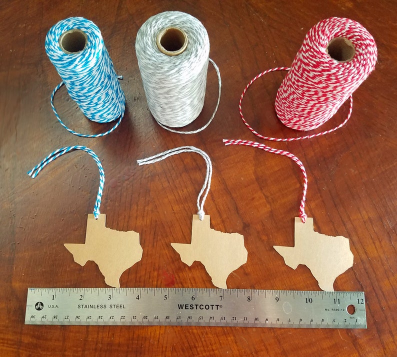 May include: Three spools of twine in blue and white, gray and white, and red and white, with matching Texas-shaped tags. A stainless steel ruler shows measurements in inches. The tags are made of brown cardboard.