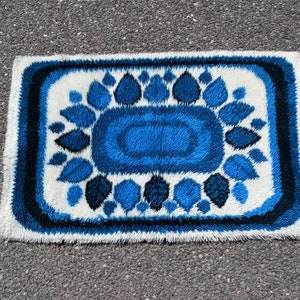 May include: A blue and white shag rug with a floral design. The rug has a white background with a blue floral pattern in the center. The rug is surrounded by a blue border.
