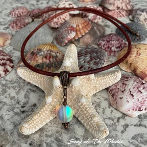 May include: A brown leather choker necklace with a copper pendant and a dichroic glass bead. The necklace rests on a white starfish, surrounded by seashells. The pendant has a decorative bail and a round, iridescent bead.
