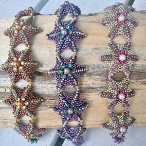 May include: Three beaded starfish bracelets in shades of brown, purple, and pink. Each bracelet features a starfish design with pearl accents. The bracelets are displayed on a wooden surface. Text on the image reads "Sheashore Bliss" and "Agnieszka Watts @ The Sea of Glass."