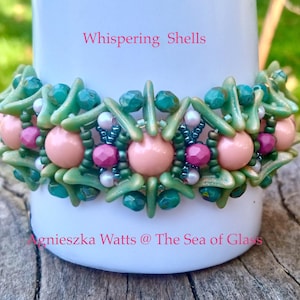 May include: A beaded bracelet with the text "Whispering Shells" and "Agnieszka Watts @ The Sea of Glass". The bracelet features green, pink, and purple beads. The bracelet is wrapped around a white cylinder.