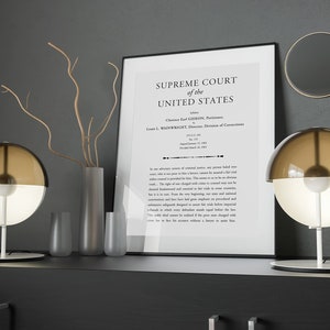 以下が含まれることがあります： 「SUPREME COURT of the UNITED STATES」というテキストと、事件名「Clarence Earl GIDEON, Petitioner, v. Wainwright, Director, Division of Corrections.」が書かれた額縁付きの文書。黒い棚の上に、丸いシェードのついた茶色と白のテーブルランプが2つ置かれている。
